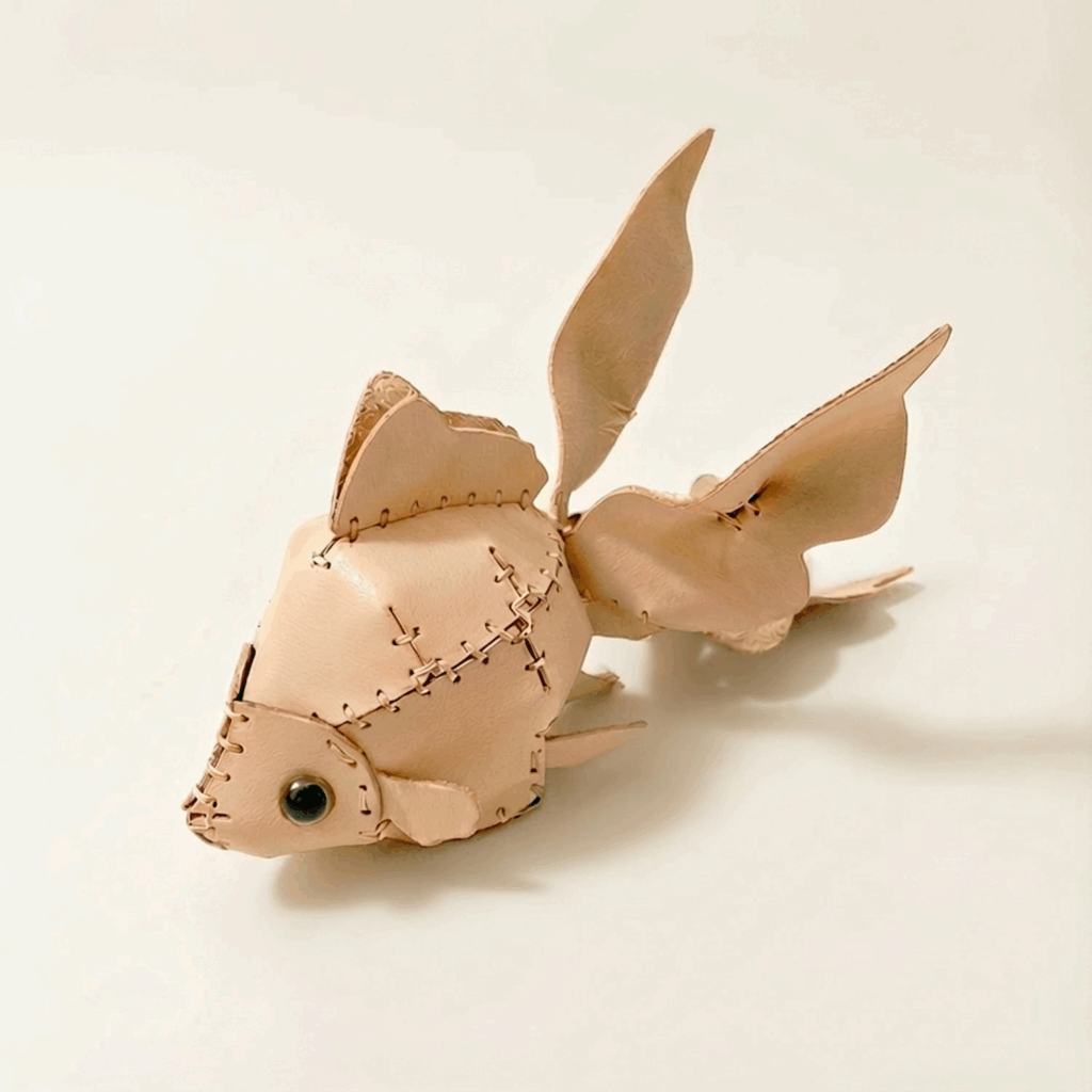 A hand-stitched natural leather goldfish with fanned tail fins and a black bead eye.